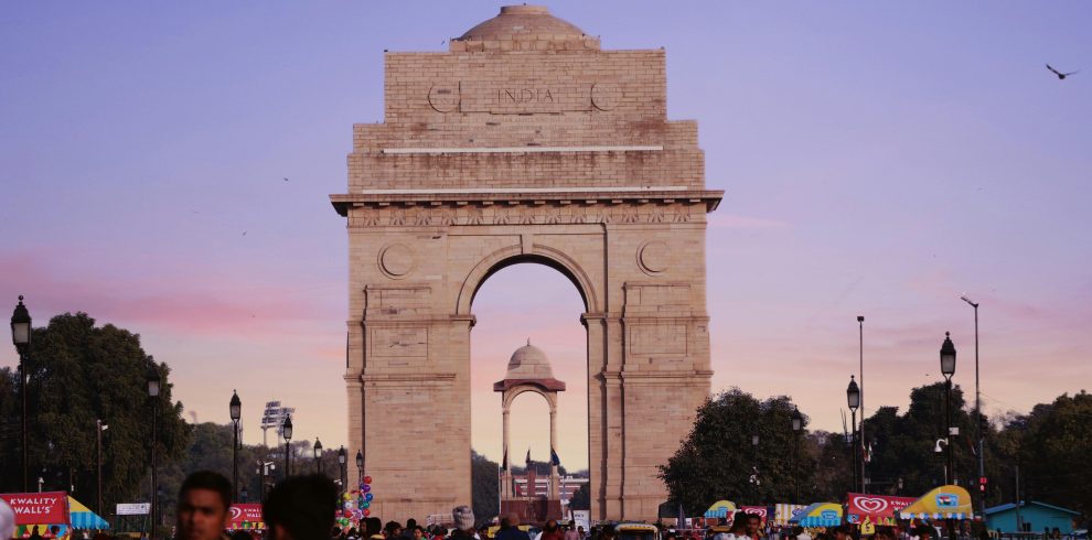 A bustling scene at India Gate with a vibrant crowd under a colorful sunset sky.